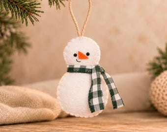 May include: A white felt snowman ornament with a stitched edge, featuring two small black bead eyes, a stitched black mouth, and a triangular orange felt nose. The snowman wears a green and white gingham fabric scarf and has a tan twine loop.