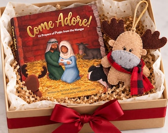May include: A gift box containing a book titled Come Adore! 12 Prayers of Praise from the Manger by Alyssa Serakos, illustrated by Saralyn Beane, and a tan felt moose ornament with dark brown antlers and a red and black buffalo plaid scarf.