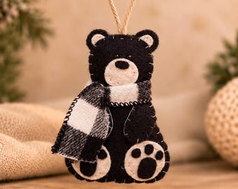 May include: A felt bear ornament in black and cream featuring a white and black buffalo plaid scarf. The bear has cream-colored paw pads and inner ears, with visible white stitching along the edges. A jute twine loop is attached to the top.