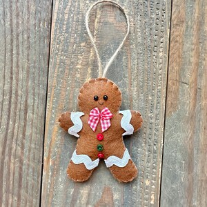 May include: A brown felt gingerbread man ornament featuring white wavy icing trim on the arms and legs, a red and white checkered bow tie, and three small buttons in red and green. The figure has two black bead eyes and a jute twine loop.