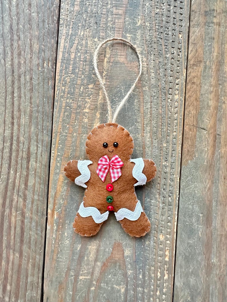 May include: A brown felt gingerbread man ornament featuring white wavy icing trim on the arms and legs, a red and white checkered bow tie, and three small buttons in red and green. The figure has two black bead eyes and a jute twine loop.