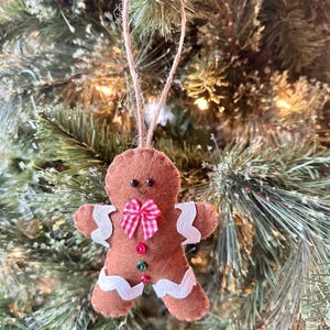 May include: A brown felt gingerbread man ornament featuring white rickrack trim on the arms and legs, a red and white gingham bow at the neck, and three small buttons in red and green down the center, suspended by a jute twine loop.