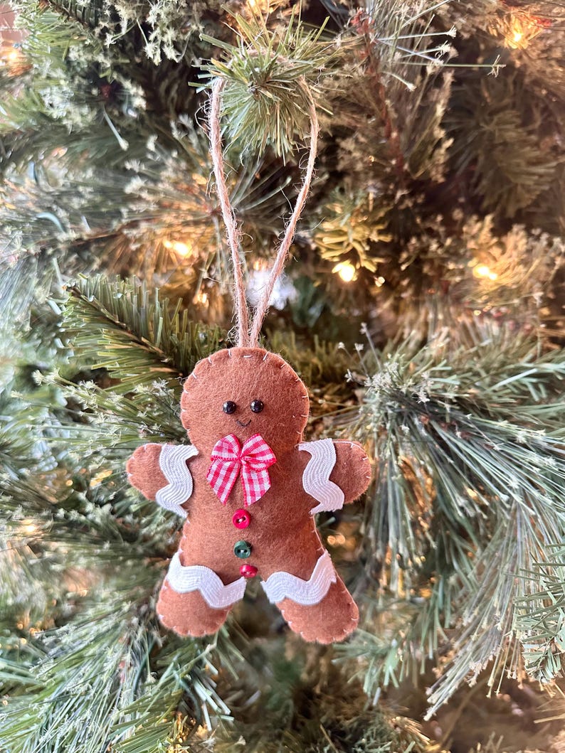 May include: A brown felt gingerbread man ornament featuring white rickrack trim on the arms and legs, a red and white gingham bow at the neck, and three small buttons in red and green down the center, suspended by a jute twine loop.