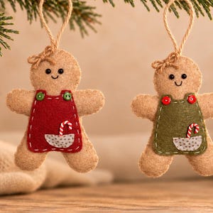 May include: Two felt gingerbread person ornaments hanging by twine loops. One wears a red apron with green buttons and a pocket containing a candy cane, the other wears a green apron with red buttons and a pocket containing a candy cane.