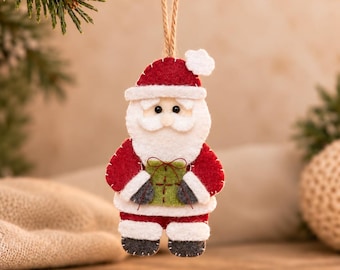 May include: A felt Santa Claus ornament featuring a red and white suit, a white beard, and a small green gift box held in the center. The figure is suspended by a thin jute twine loop and includes dark gray boots and mittens with white trim.