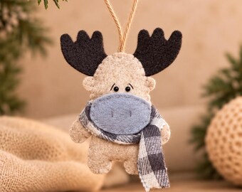 May include: A felt moose ornament in beige with dark brown antlers, a light blue muzzle, and a black and white buffalo plaid scarf. The figure features small black bead eyes and is suspended by a thin jute twine loop for hanging.