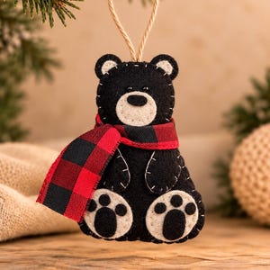 May include: A black felt bear ornament with a cream-colored muzzle and paw pads. The bear features white decorative stitching, small black eyes, and a red and black buffalo plaid fabric scarf. A jute twine loop is attached to the top for hanging.