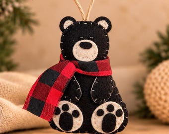 May include: A black felt bear ornament with a cream-colored muzzle and paw pads. The bear features white decorative stitching, small black eyes, and a red and black buffalo plaid fabric scarf. A jute twine loop is attached to the top for hanging.