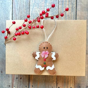 May include: A brown felt gingerbread man ornament with white wavy icing accents, a red and white checkered bow, and two small buttons. The ornament features black bead eyes and a stitched smile, hanging from a jute string near a red berry sprig.