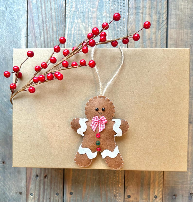 May include: A brown felt gingerbread man ornament with white wavy icing accents, a red and white checkered bow, and two small buttons. The ornament features black bead eyes and a stitched smile, hanging from a jute string near a red berry sprig.