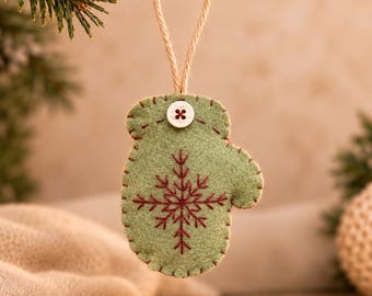 May include: A sage green felt mitten ornament featuring a dark red embroidered snowflake design on the front. The mitten is accented with a small white button at the top and hangs from a simple tan twine loop for display purposes.