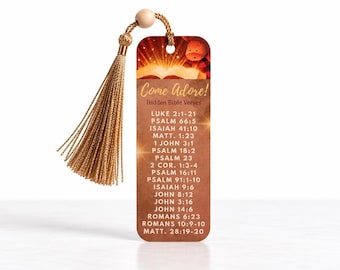 May include: A rectangular bookmark with a gold tassel and a wooden bead. The design features a glowing open book graphic at the top, the text "Come Adore! Hidden Bible Verses," and a list of Bible citations in white font on a brown background.
