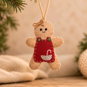May include: A tan felt gingerbread person ornament featuring a red apron with two green buttons, a small gray pocket, and a candy cane graphic. The figure has a stitched face, a small bow on its head, and a jute twine loop for hanging.