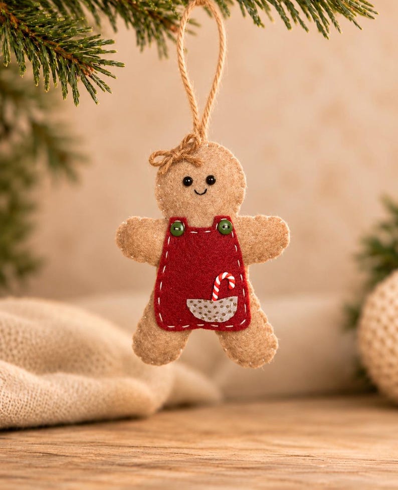 May include: A tan felt gingerbread person ornament featuring a red apron with two green buttons, a small gray pocket, and a candy cane graphic. The figure has a stitched face, a small bow on its head, and a jute twine loop for hanging.