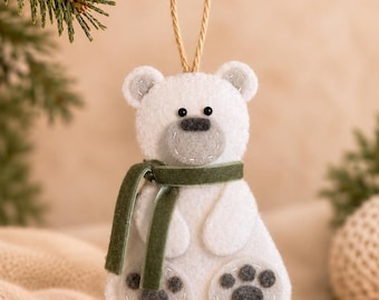 May include: A white felt polar bear ornament with a gray snout, black bead eyes, and a dark green velvet ribbon scarf. The bear features gray felt paw pads on its feet and is suspended by a thin tan twine loop for hanging on a tree branch.