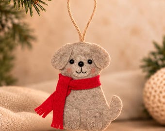 May include: A gray felt dog ornament featuring a bright red scarf, black bead eyes, and a stitched nose and mouth. The ornament is suspended by a thin tan twine loop, showcasing detailed stitching on the paws and a small upright tail.