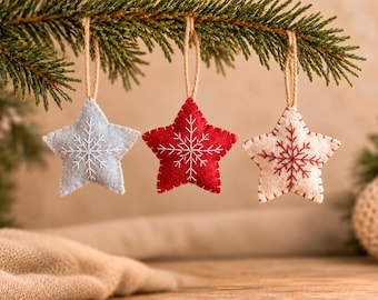 May include: Three star-shaped felt ornaments hanging from a pine branch. The left ornament is light blue with white stitching, the center is red with white stitching, and the right is white with red stitching, each featuring a snowflake design.