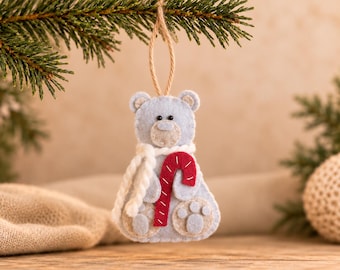 May include: A light blue felt bear ornament featuring a white yarn scarf and a red and white striped candy cane held in its paws. The bear has a beige muzzle, small black bead eyes, and beige paw pads on its feet, suspended by a jute twine loop.