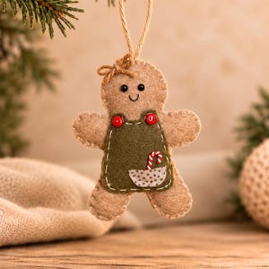 May include: A tan felt gingerbread person ornament with a small brown bow on its head. It wears a dark green felt apron with two red buttons, a small pocket, and a miniature candy cane tucked inside. The ornament hangs from a jute string.