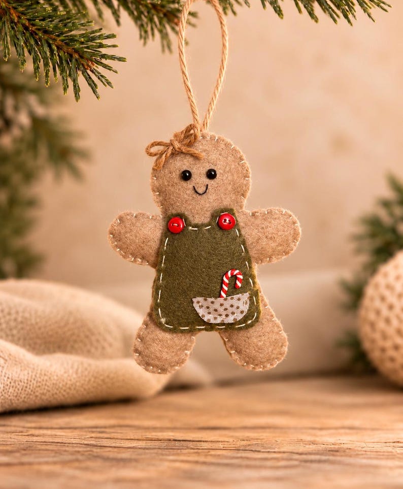 May include: A tan felt gingerbread person ornament with a small brown bow on its head. It wears a dark green felt apron with two red buttons, a small pocket, and a miniature candy cane tucked inside. The ornament hangs from a jute string.