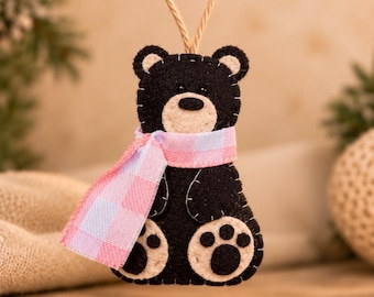May include: A black felt bear ornament with a beige muzzle and paw pads, featuring visible whip-stitch detailing. The bear wears a pink and light blue checkered fabric scarf and hangs from a thin, natural-colored jute twine loop.