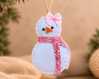 May include: A light blue felt snowman ornament featuring a small pink satin bow on its head, a bright orange felt carrot nose, black bead eyes, and a stitched mouth. The snowman wears a long, sparkly pink glitter scarf and hangs from a jute twine loop.