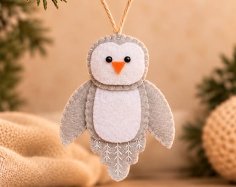 May include: A felt owl ornament featuring a gray and white body with a bright orange triangular beak and two small black bead eyes. The owl has stitched details on its wings and tail, and it hangs from a simple tan twine loop.