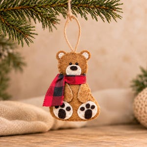 May include: A brown felt bear ornament featuring a red and black buffalo plaid scarf, white muzzle, and black stitched nose and eyes. The bear has white felt paw pads with black stitched details and hangs from a thin jute twine loop.