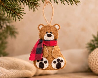 May include: A brown felt bear ornament featuring a red and black buffalo plaid scarf, white muzzle, and black stitched nose and eyes. The bear has white felt paw pads with black stitched details and hangs from a thin jute twine loop.