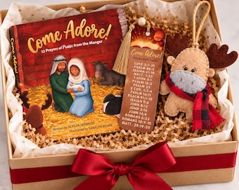 May include: A gift box containing a book titled Come Adore! 12 Prayers of Praise from the Manger, a bookmark listing Bible verses, and a felt moose ornament with a red and black plaid scarf, all tied with a large red satin ribbon.