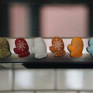 May include: A row of six felt mittens in various colors, including green, red, white, brown, orange, and blue. Each mitten features a white snowflake design. The mittens are likely decorative items.