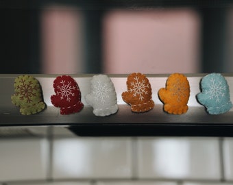 May include: A row of six felt mittens in various colors, including green, red, white, brown, orange, and blue. Each mitten features a white snowflake design. The mittens are likely decorative items.