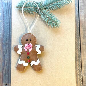May include: A brown felt gingerbread man ornament featuring white wavy icing details, a red and white checkered bow, two small buttons, and black bead eyes. The ornament hangs from a jute string near a sprig of green pine needles.