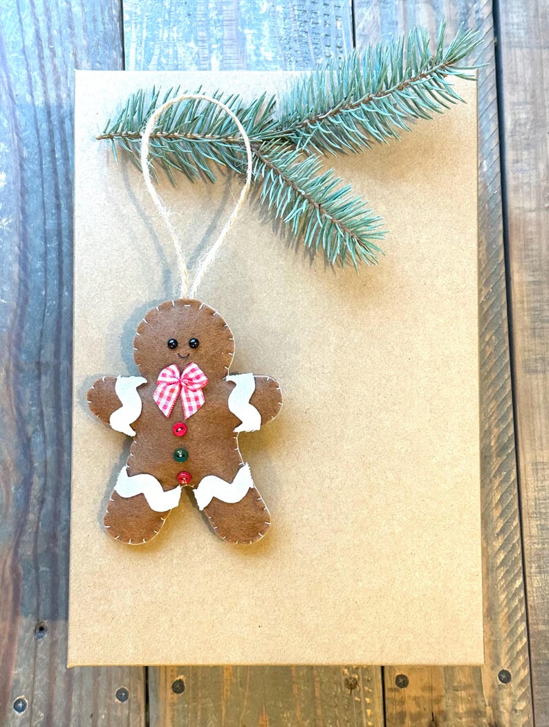 May include: A brown felt gingerbread man ornament featuring white wavy icing details, a red and white checkered bow, two small buttons, and black bead eyes. The ornament hangs from a jute string near a sprig of green pine needles.