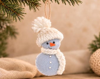 May include: A felt snowman ornament in light blue with a white knitted pom-pom hat and a matching white braided scarf. The snowman features a small orange felt carrot nose, black stitched eyes, a curved mouth, and three white buttons.