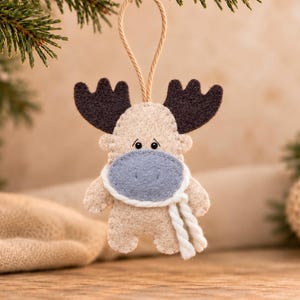 May include: A felt moose ornament in beige with dark brown antlers, a light blue muzzle, and a white twisted yarn scarf. The ornament features small black bead eyes and is suspended by a thin tan cord for hanging, showcasing detailed stitching.