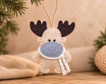 May include: A felt moose ornament in beige with dark brown antlers, a light blue muzzle, and a white twisted yarn scarf. The ornament features small black bead eyes and is suspended by a thin tan cord for hanging, showcasing detailed stitching.