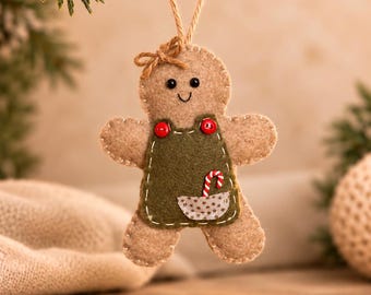 May include: A tan felt gingerbread person ornament with a small brown bow on its head. It wears a dark green felt apron with two red buttons, a small pocket, and a miniature candy cane tucked inside. The ornament hangs from a jute string.