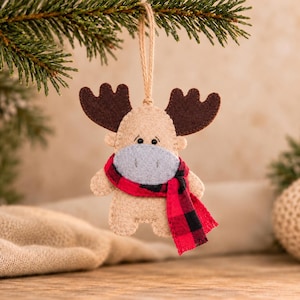 May include: A felt moose ornament in beige with dark brown antlers, a light gray muzzle, and a red and black buffalo plaid scarf. The figure features small black bead eyes and is suspended by a thin jute twine loop for hanging purposes.