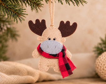 May include: A felt moose ornament in beige with dark brown antlers, a light gray muzzle, and a red and black buffalo plaid scarf. The figure features small black bead eyes and is suspended by a thin jute twine loop for hanging purposes.