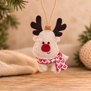 May include: A felt reindeer ornament featuring a light beige body, dark brown antlers, and a bright red nose. The reindeer wears a red and white patterned scarf and hangs from a tan twine loop. The ornament has stitched edges and small black bead eyes.