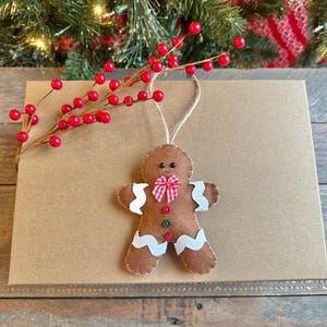 May include: A brown felt gingerbread man ornament featuring white rickrack trim on the arms and legs, a red and white gingham bow tie, and two small buttons in red and green. The ornament hangs from a jute twine loop.