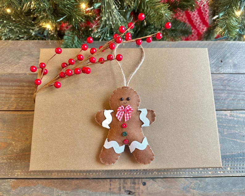 May include: A brown felt gingerbread man ornament featuring white rickrack trim on the arms and legs, a red and white gingham bow tie, and two small buttons in red and green. The ornament hangs from a jute twine loop.