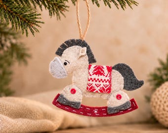 May include: A felt rocking horse ornament in beige and dark gray with a red and white patterned saddle blanket. The horse features small red buttons at the joints and a red rocker base with white stitching details, suspended by a jute twine loop.