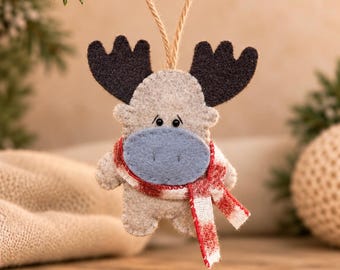 May include: A felt moose ornament in light gray with dark gray antlers and a blue snout. The moose wears a red and white plaid scarf and hangs from a tan twine loop. The figure features simple black stitched eyes and nose details.