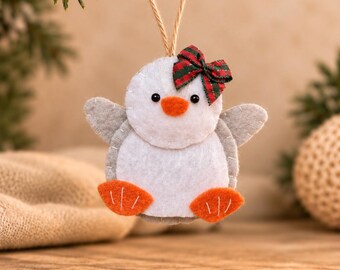 May include: A felt penguin ornament featuring a white face and belly, gray wings, and bright orange feet and beak. The penguin has two small black bead eyes and a small red and green plaid ribbon bow attached to the top of its head. A thin jute twine loop is attached for hanging.