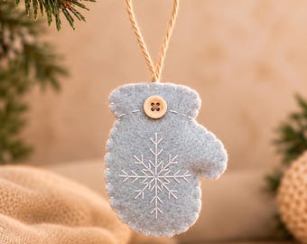 May include: A light blue felt mitten ornament featuring white embroidered snowflake detailing and a small wooden button at the top. The mitten is suspended by a thin jute twine loop, showcasing visible white blanket stitching along the edges.
