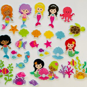 May include: A collection of colorful mermaid and sea creature cutouts. The mermaids have various hair colors and rainbow-colored tails. Other sea creatures include an octopus, turtle, starfish, fish, and crab. The cutouts are designed for crafting or decoration.