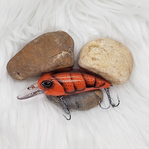 May include: A vibrant orange and black fishing lure with a clear plastic lip and two treble hooks. The lure is designed to mimic a crawfish, resting on a bed of white faux fur with two stones.