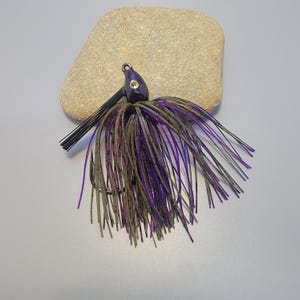 May include: A purple and black fishing jig with a hook. The jig has a dark purple head, black bristles, and a skirt of olive green and purple strands. The fishing lure is resting on a light brown stone.